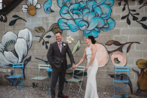 bride and groom laughing and holding hands in front of a flower mural in detroit