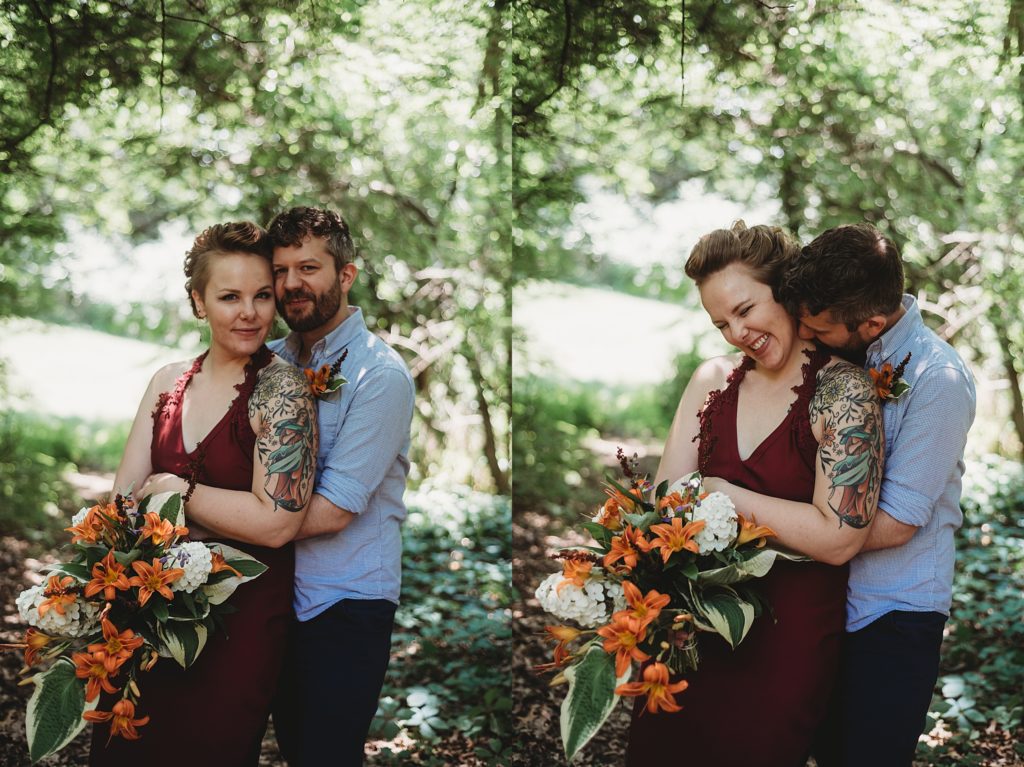 bride in a maroon dress with orange flowers and tattoos and groom in a blue shirt smiling and laughing in the woods