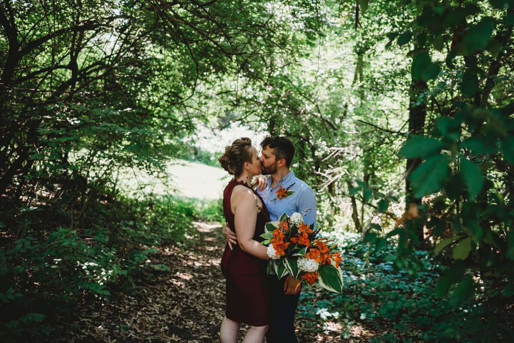 bride in a maroon dress with orange flowers and tattoos and groom in a blue shirt smiling and laughing in the woods