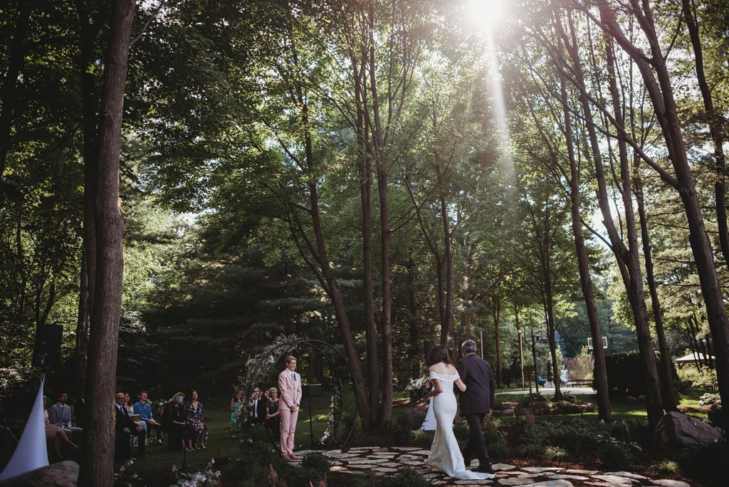 bride in a pink suit crying as she sees her wife walking down the aisle towards her in the woods