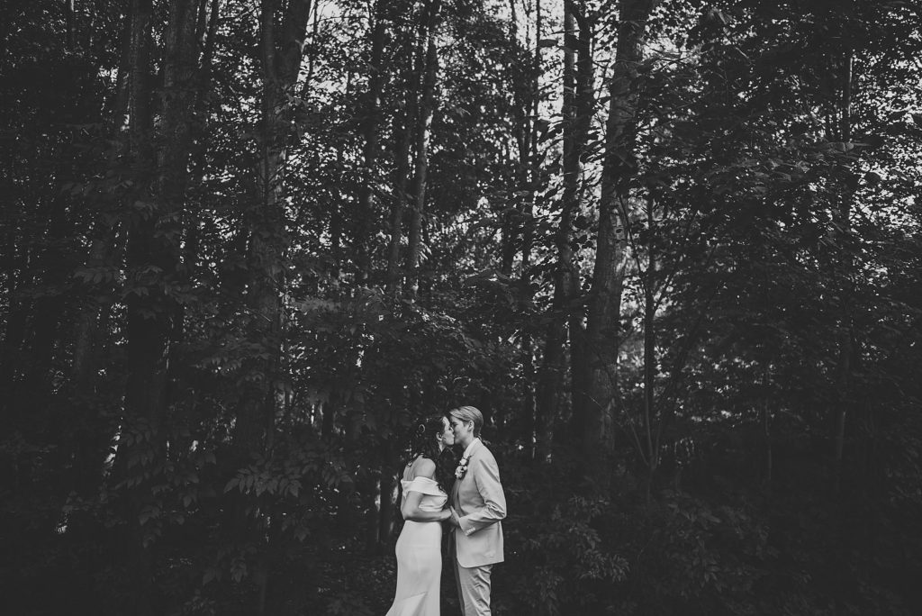 bride in pink suit and bride in a white dress cuddling and kissing her forehead in the woods