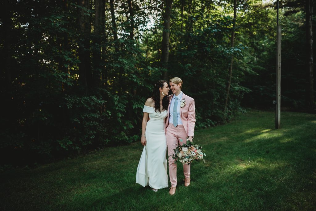 bride in a pink suit and bride in a white dress walking and laughing while holding pink and white wedding flowers