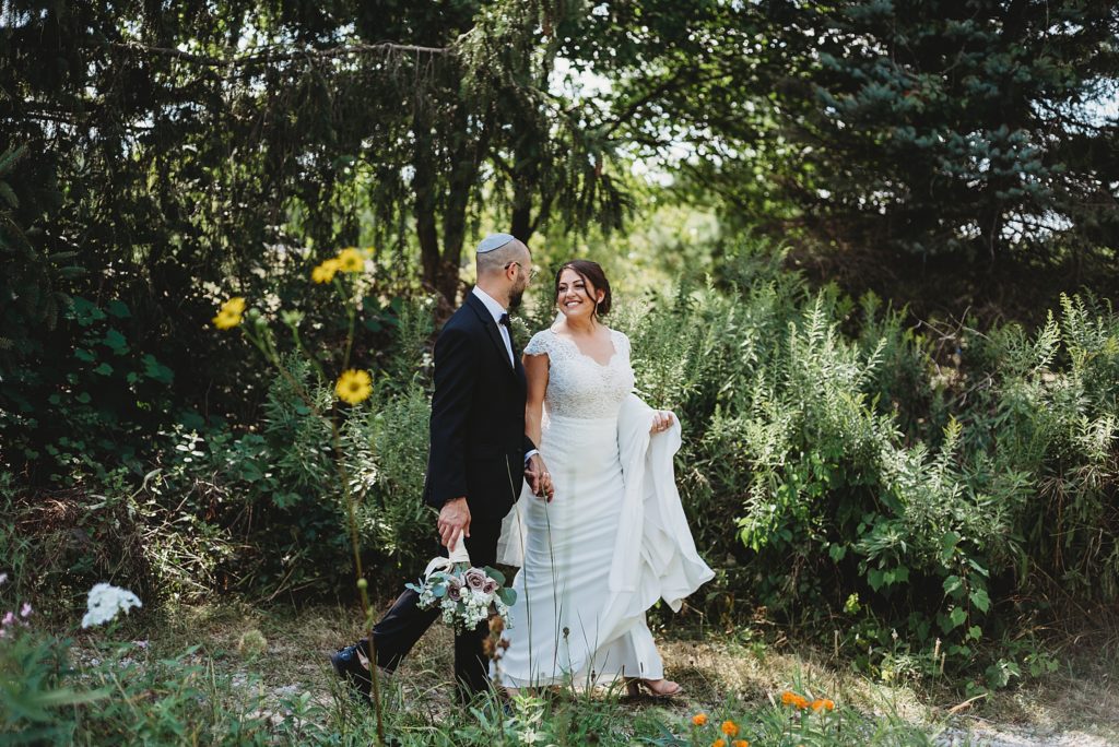 groom in a Yamaka and bride in a white lace dress smiling and laughing with each other in the woods. Bride holding a pink rose bouquet