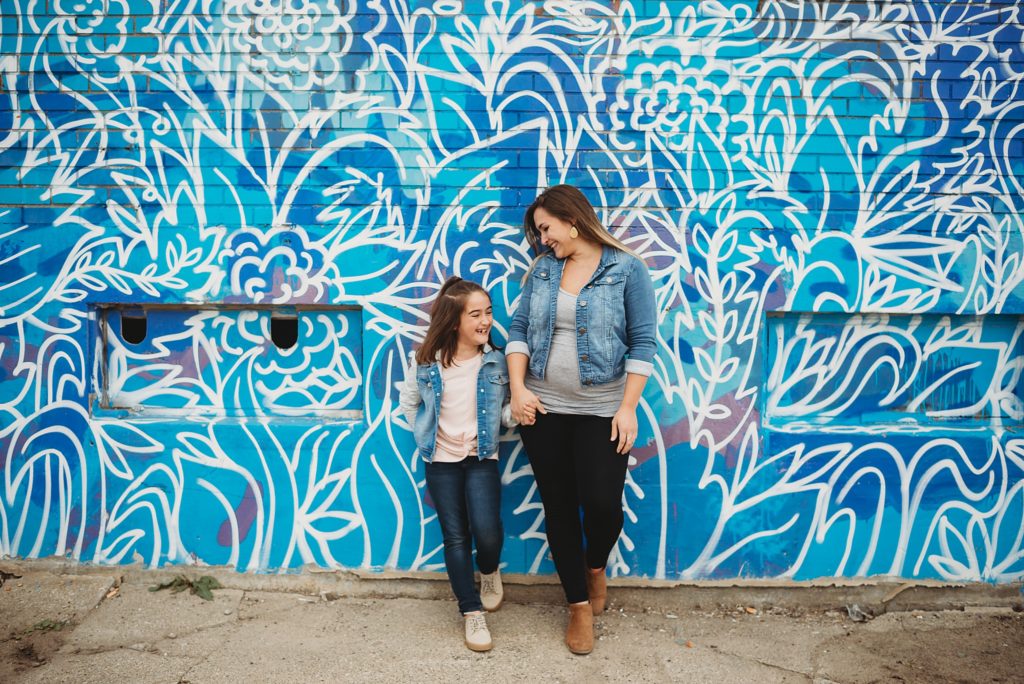 pregnant mom and daughter smiling at each other standing against a graffiti wall 