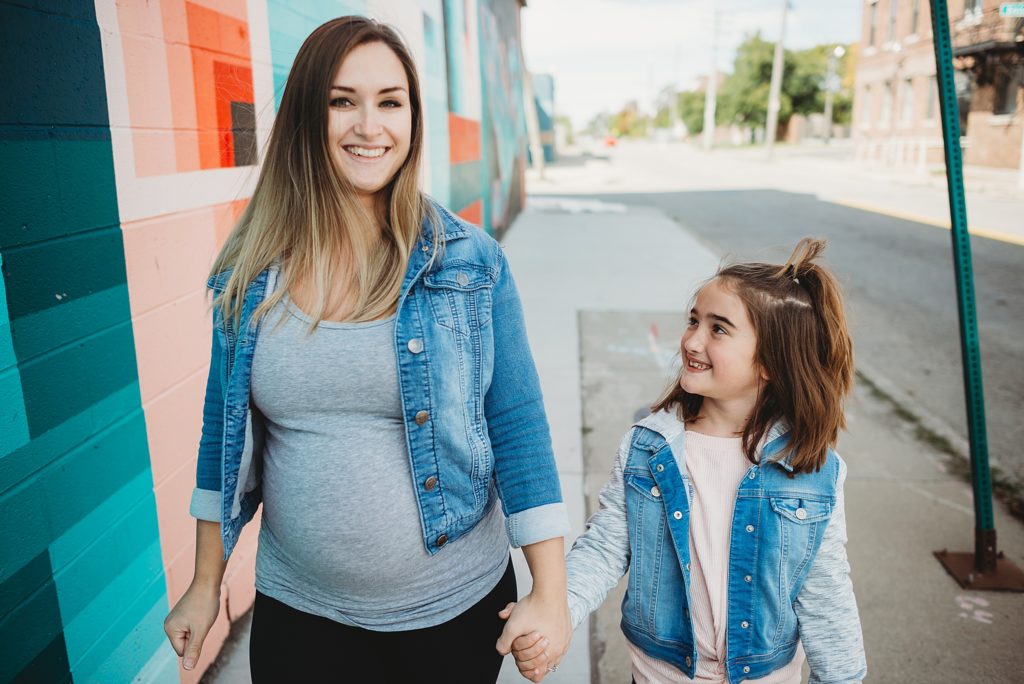 pregnant mom and daughter holding hands and walking down the sidewalk laughing and smiling 