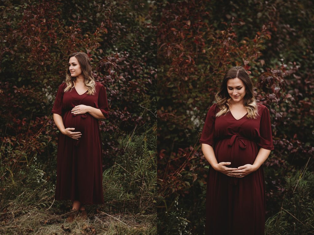 pregnant mom in a maroon dress holding her belly smiling with the fall leaves behind her