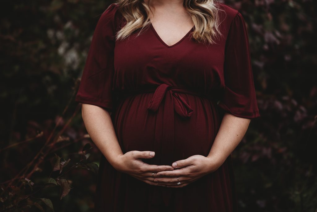 pregnant mom in a maroon dress holding her belly smiling with the fall leaves behind her