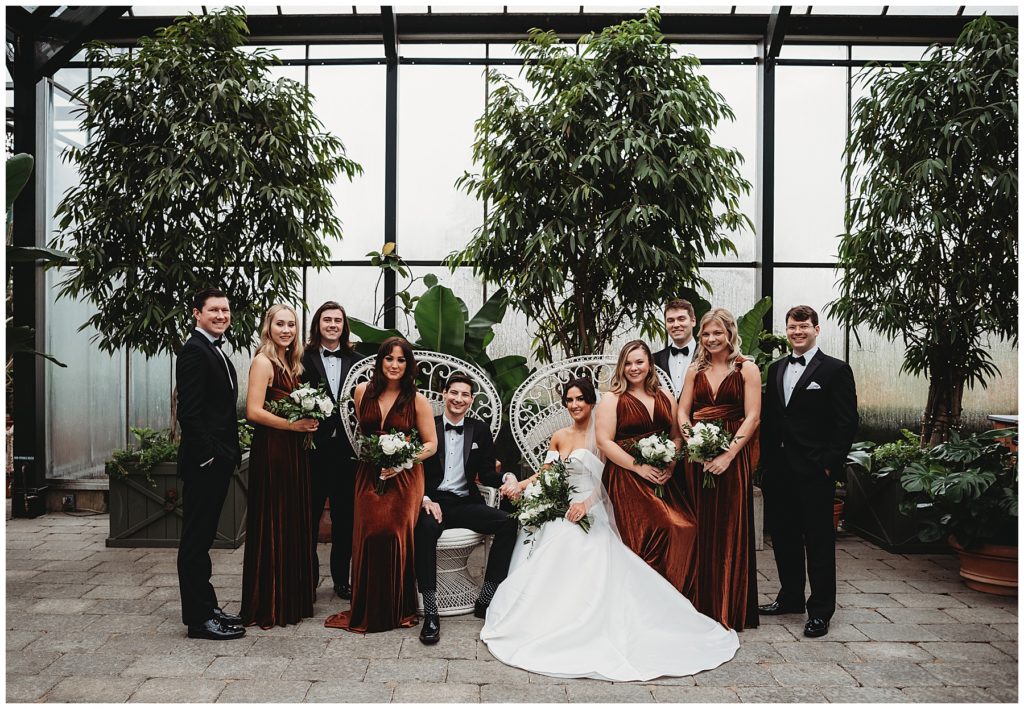 wedding party posing and smiling around the bride and groom sitting in peacock chairs. Bridesmaids wearing coper velvet wrap dresses and groomsmen in formal black suits. Holding white floral bouquets.